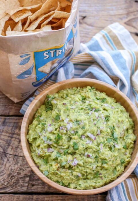 "Bowl of green chile guacamole with tortilla chips and lime wedges on rustic table"